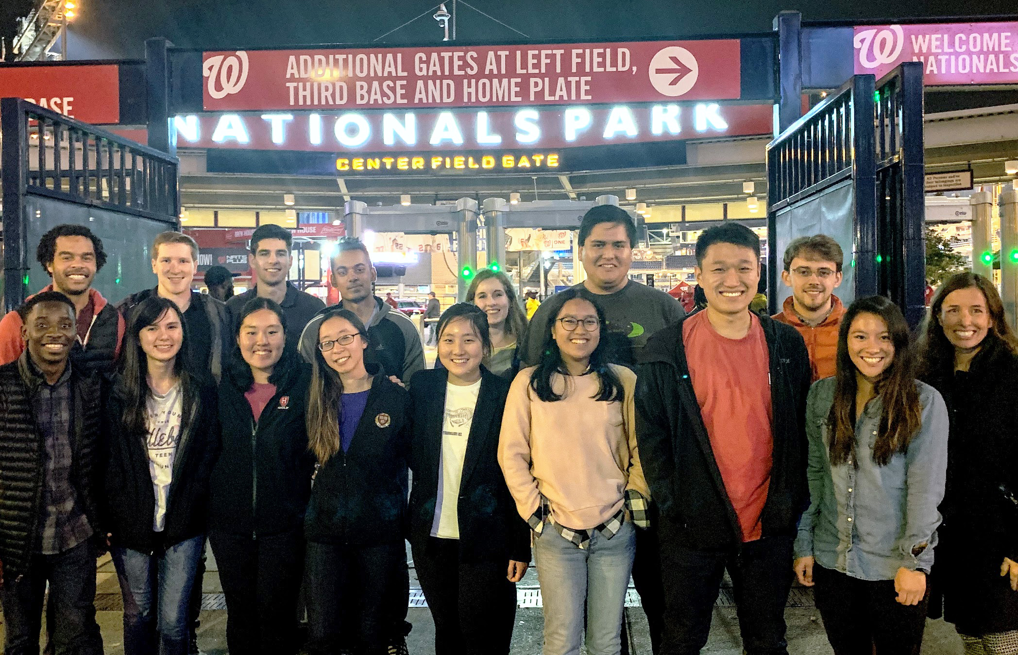 The whole group wears warm, casual clothes, standing outside bright lights advertising the Nationals baseball park at night. The group stayed nearby, but the Nationals were playing an away game in Houston.