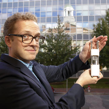 Christoffer wears a navy suit over a bright blue and gray checkered shirt. He looks sideways at the camera as he presents MagLev, a thermos-sized tool with white blocks held together with thick screws and a clear