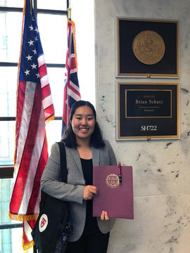 Ji Hae Lee stands in front of the American and Hawaii flags, wearing a gray suit jacket over a black shirt and slacks. She holds a Harvard University folder up for the camera.