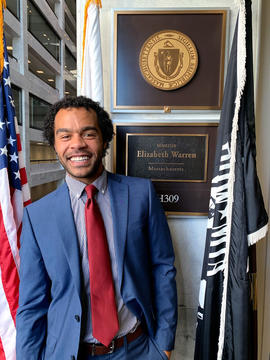 Zach Zinsli wears a blue suit over a checkered blue and white shirt and a red tie. He stands next to American and Massachusetts flags and a mahogany plaque with Senator Elizabeth Warren's name and state.