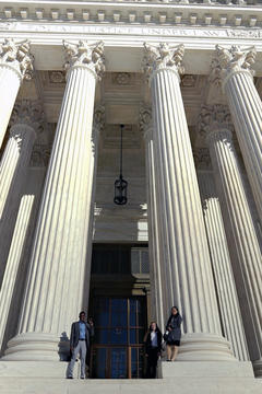 At the base of huge, ornate white columns, three students pose: Augustine, standing beneath a column to the left of the building entrance, makes a peace sign while Amanda and Christina stand to the right, Christina on top of the column's wide base.