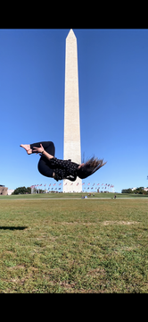 In mid-flip in front of the Washington monument, Michelle hangs perpendicular to the ground, her hair whipping toward the sky. She is barefoot, wearing black pants and a patterned blouse.