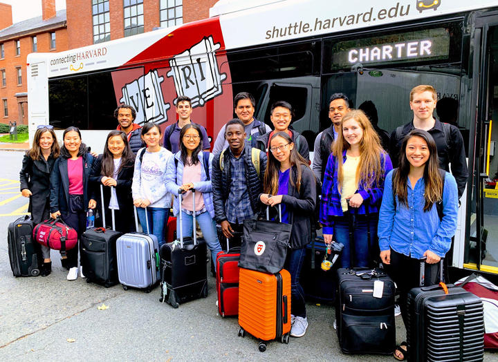 A group of twelve students and three instructors stand with their suitcases in front of a Harvard shuttle