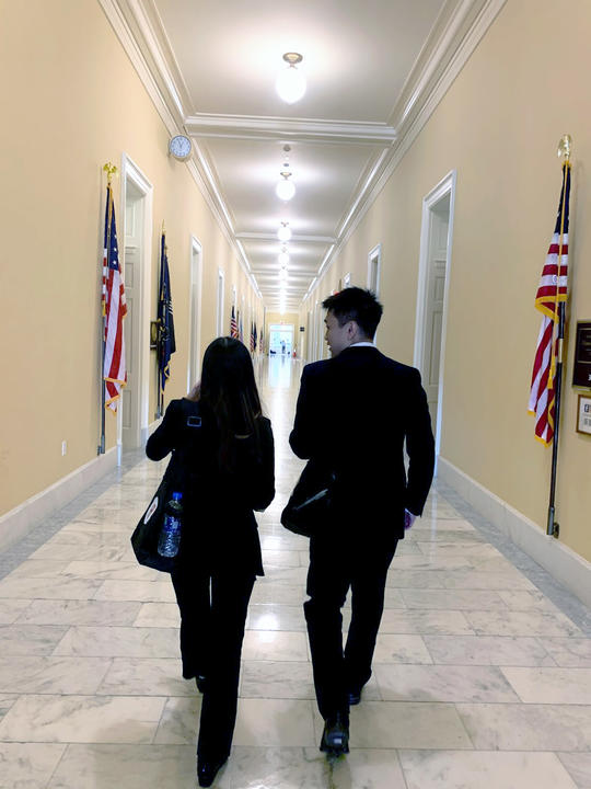 Both in black suits, Stephanie and Eugene walk away from the camera, down a hallway with white marble floors, soft yellow walls, and American flags stationed against the walls.
