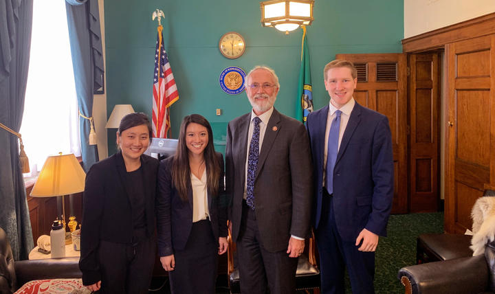 Rep. Dan Newhouse wears a white beard, glasses, gray suit and tie with small flowers in various blues. The students wear dark suits, standing to his left and right.
