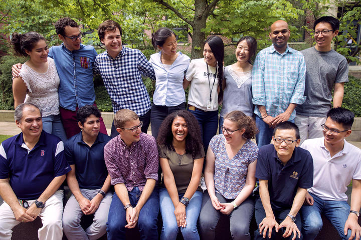 In front of green leaves and a brick building, Christina Woo stands in a row of her lab members with another row seated in front. The shot captures an informal moment: Woo and her diverse group of lab members smile and laugh, looking around at each other