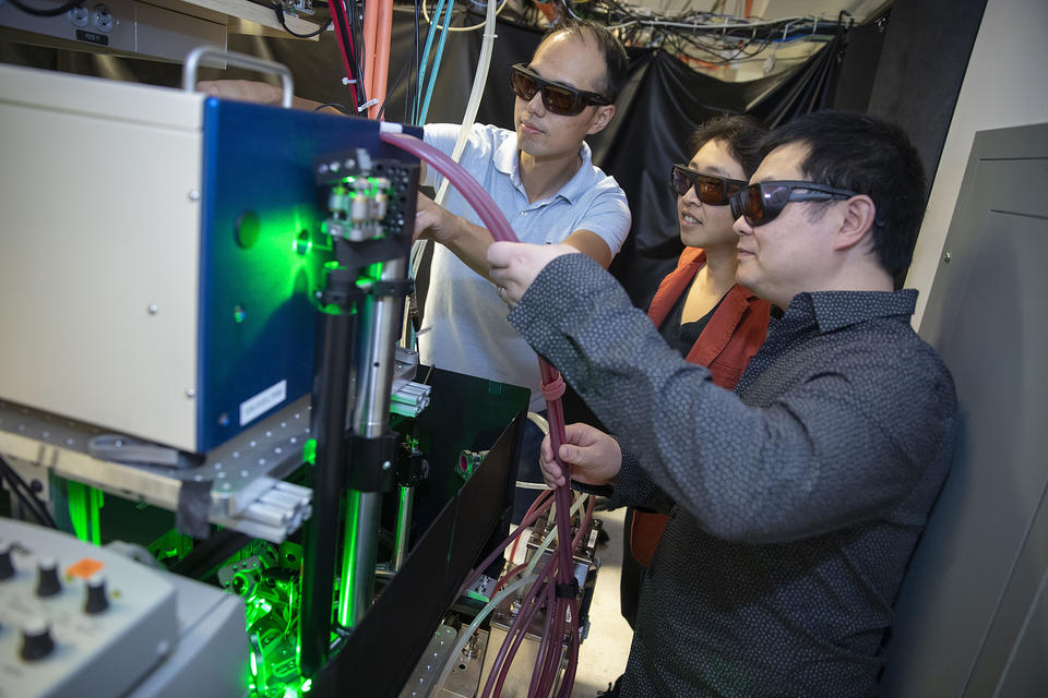 A photo of Kang-Kuen Ni, Ming-Guang, and Yu Liu standing at their wire-filled machine in the lab