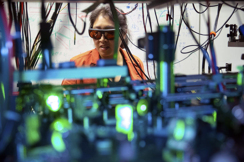 A photo of Kang-Kuen Ni standing behind machines that glow green and tangled wires in her lab