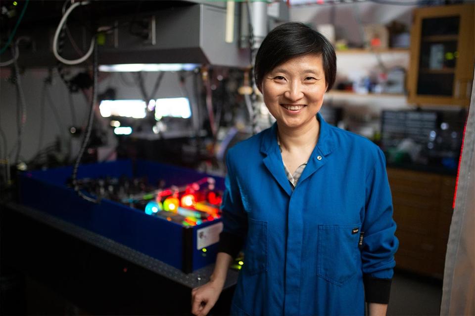 A photo of Xiaowei Zhuang smiling while she wears a blue lab coat and stands in her lab