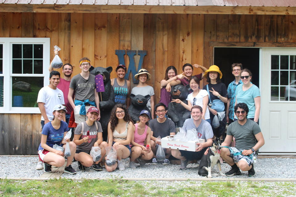 A group photo of Woo lab members on a lab outing: They're posing in front of a wooden cabin and holding paintballs