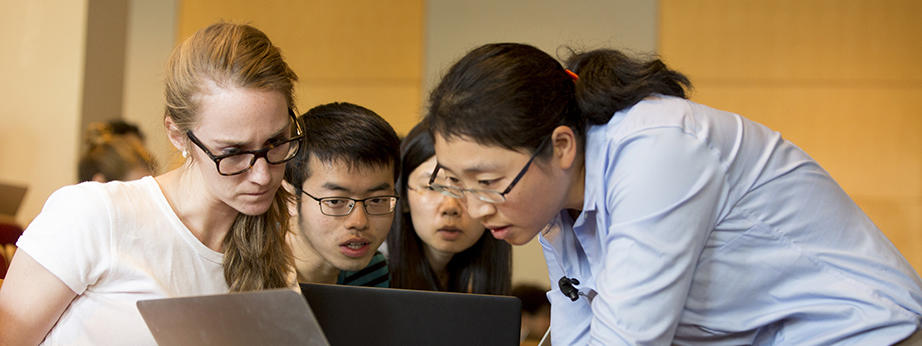 Assistant Professor Kang-Kuen Ni, in a light blue button up, her hair tied back in a ponytail, leans over to look at a female student's laptop as two other students look on in the background