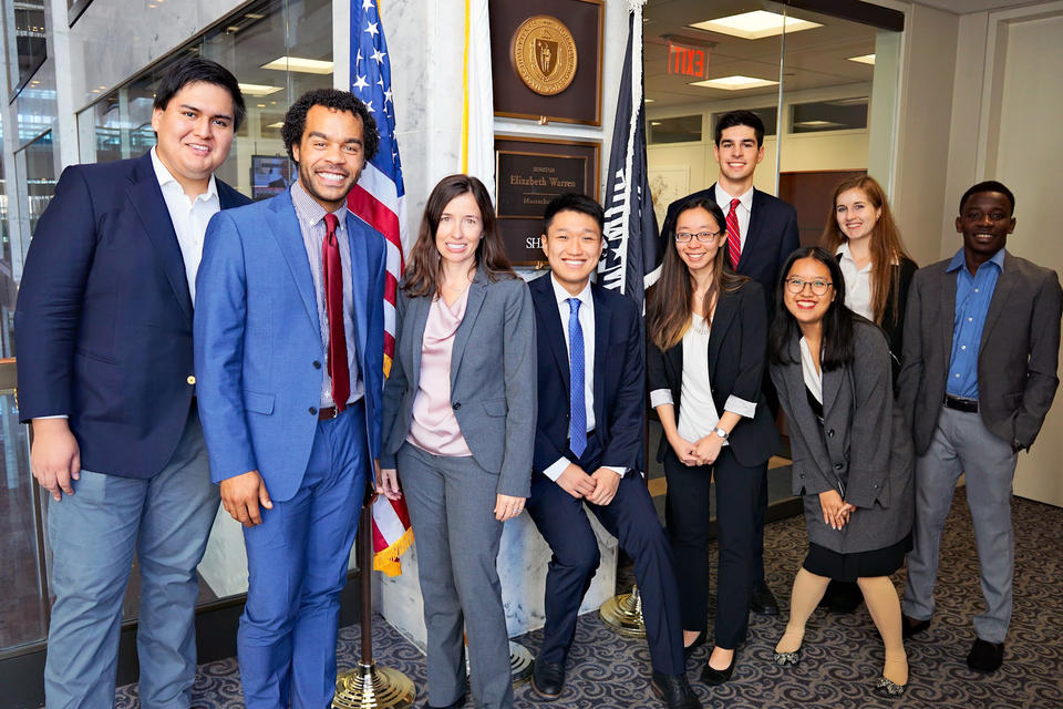 Nine members of the advocacy trip pose in front of the plaque outside Senator Warren's office. They wear dark suits for their meetings on Capitol Hill.